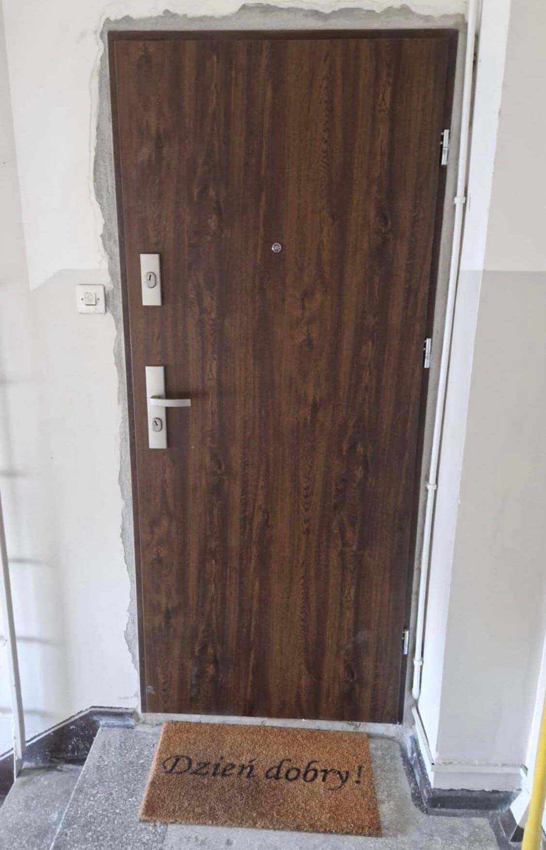 Brązowe drzwi zewnętrzne z wizjerem i srebrną klamką, zamontowane w surowym otworze ściany, z wycieraczką z napisem 'Dzień dobry!' na progu.