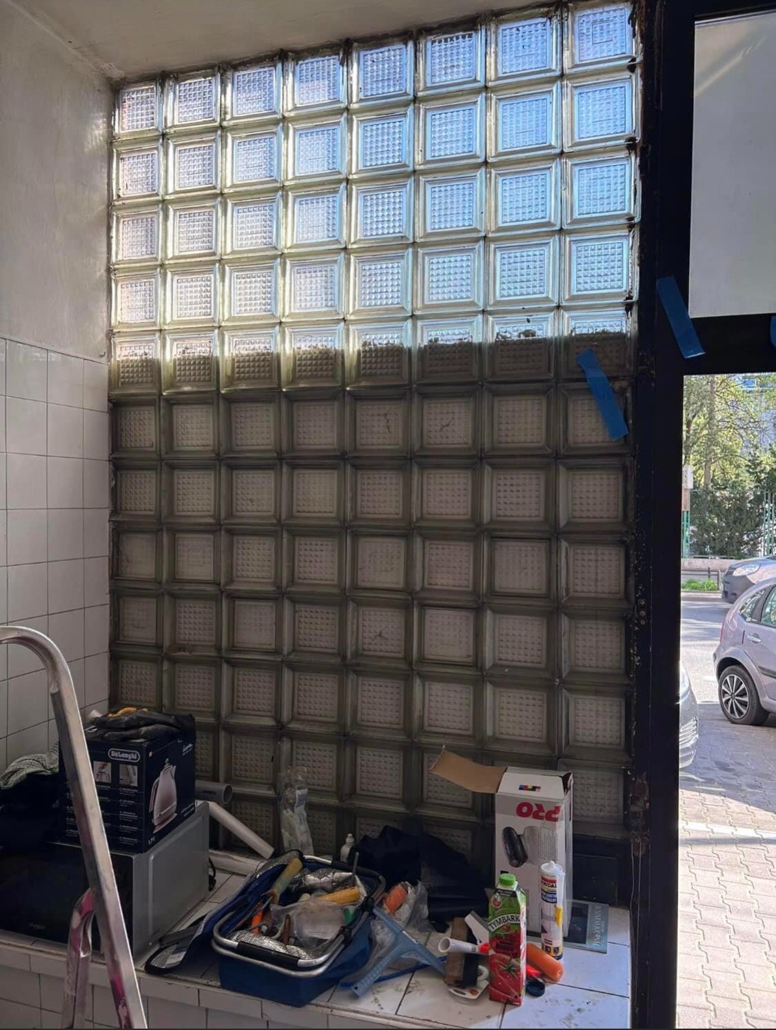 Ściana z luksferów w trakcie montażu, widoczne narzędzia i materiały budowlane, częściowo wykończona, z widokiem na ulicę przez otwarte drzwi