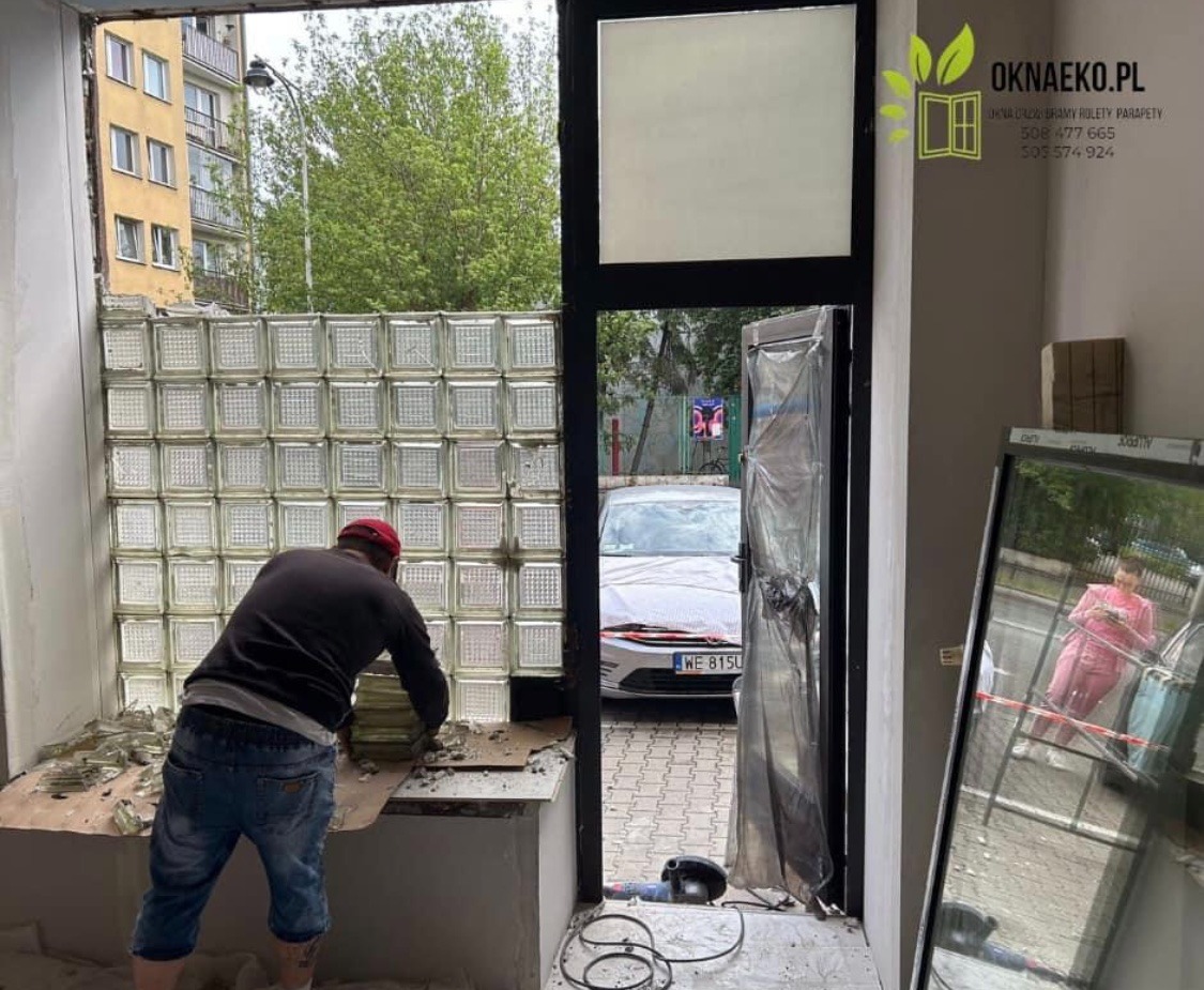 Pracownik w czerwonej czapce demontuje ścianę z luksferów, przygotowując otwór na nowe okno. Na zewnątrz widoczny zaparkowany samochód i budynek mieszkalny.