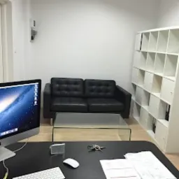 Wnętrze biura z czarną sk&oacute;rzaną sofą, szklanym stolikiem kawowym, białym regałem z przegr&oacute;dkami i komputerem Apple iMac na czarnym biurku. Na biurku leżą klucze, myszka i dokumenty.