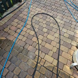 Wysokociśnieniowi Marcin Szczepanik - Mokre kostki brukowe po czyszczeniu, widoczny fragment urządzenia czyszczącego, niebieski wąż doprowadzający wodę i czarny kabel zasilający.