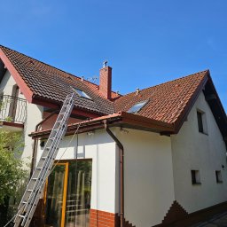 WYSOKOCIŚNIENIOWI - Dwupiętrowy dom z czerwonym dachem widziany z dołu, z aluminiową drabiną opartą o rynnę, wskazującą na prace konserwacyjne lub instalacyjne.
