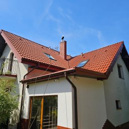 WYSOKOCIŚNIENIOWI - Dwupiętrowy dom z czerwonym dachem i białymi ścianami, z widocznymi rynnami i oknami dachowymi, na kominie siedzi bocian, wzdłuż ściany rozciągnięty przewód.