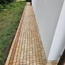 Ułożona z kostki brukowej ścieżka wzdłuż jasnej ściany budynku z widoczną rynną spustową i kratką odpływową, obok trawnik.
