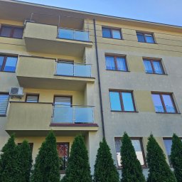 Wysokociśnieniowi Marcin Szczepanik - Wielopiętrowy budynek z beżową elewacją, balkonami z szklanymi balustradami i rzędem iglaków u podstawy.