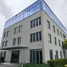 Wysokociśnieniowi Marcin Szczepanik - Nowoczesny, trzypiętrowy budynek biurowy z przeszkloną nadbudówką, widoczny z poziomu chodnika przed wejściem, gdzie widać dwie osoby wchodzące do budynku.