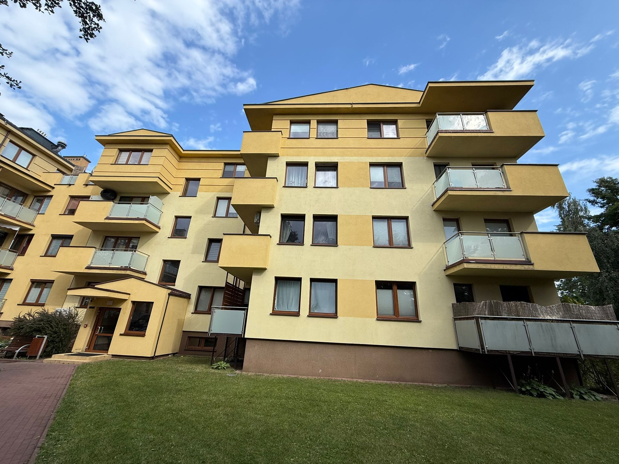 Żółty budynek wielorodzinny z balkonami i brązowymi ramami okiennymi, widziany z perspektywy trawnika w słoneczny dzień. Widoczne błękitne niebo z chmurami.