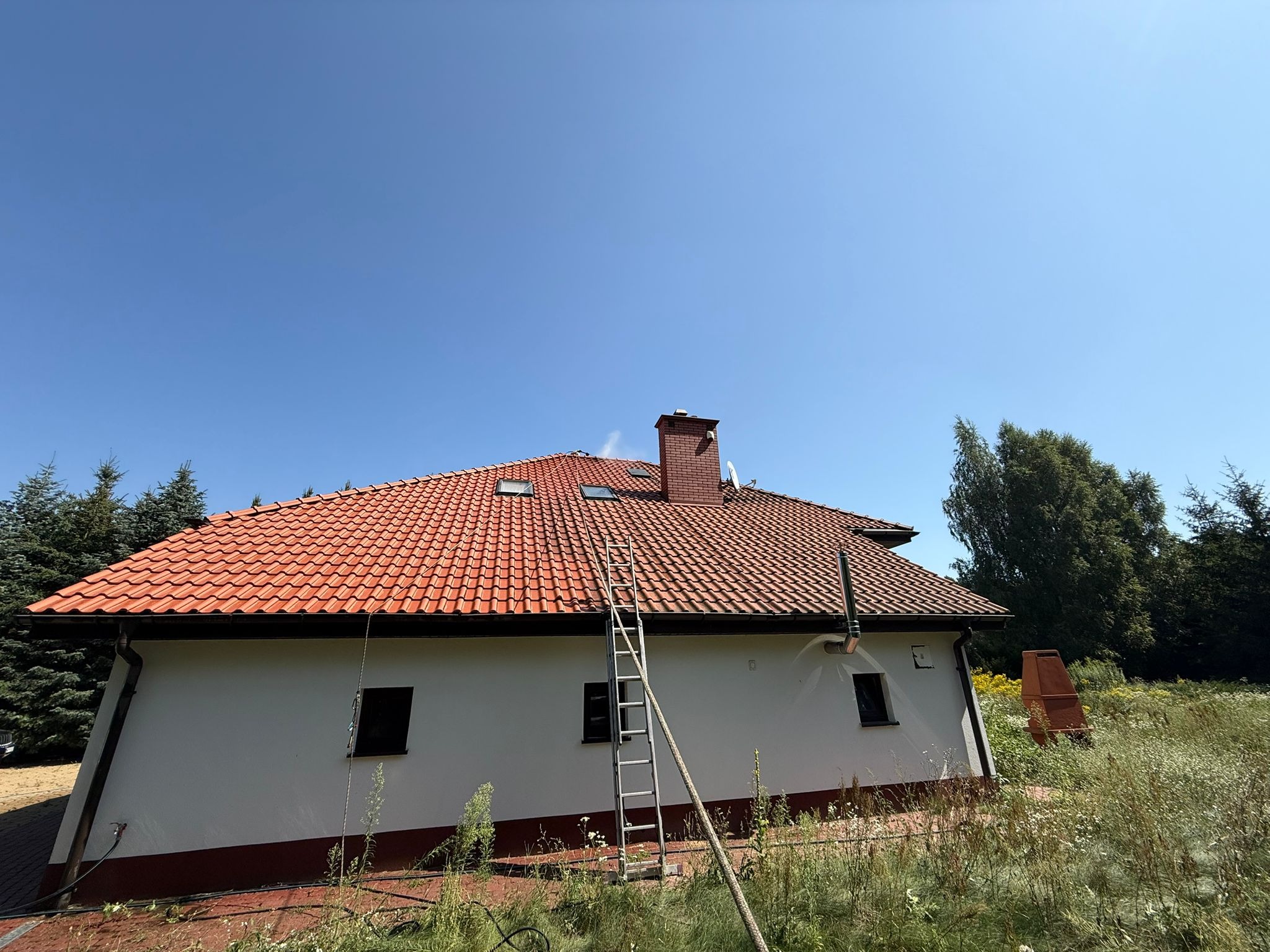 Dom jednorodzinny z czerwoną dachówką, drabina oparta o dach, prawdopodobnie w trakcie czyszczenia. Błękitne niebo w tle, widoczne drzewa i trawa.