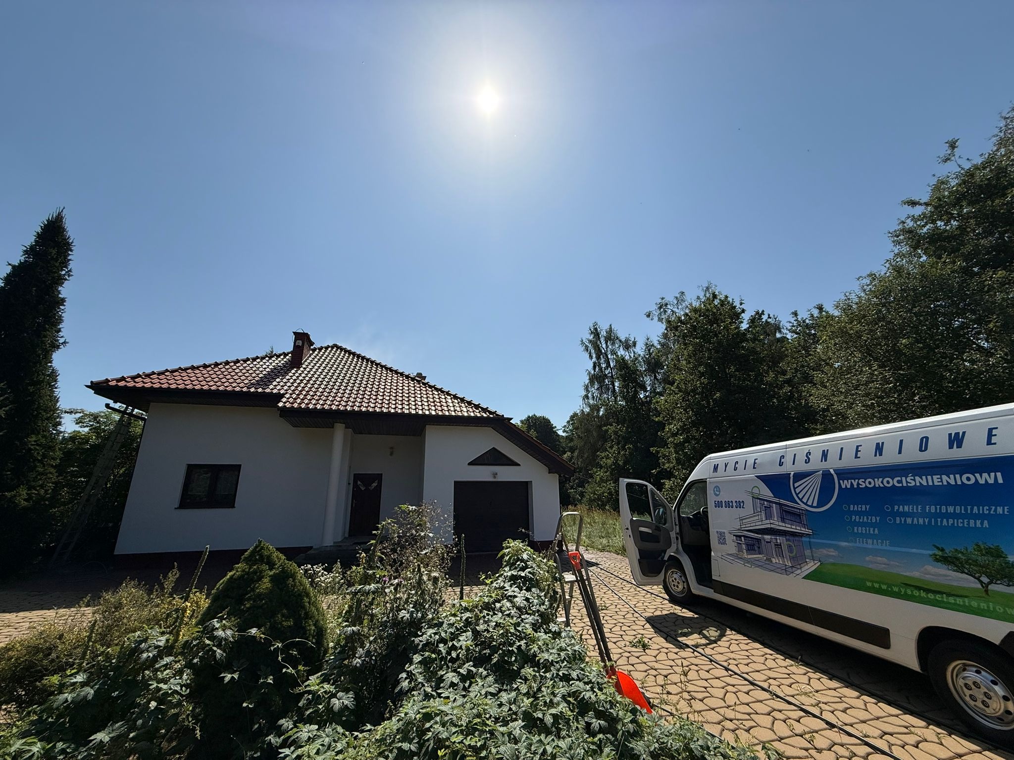 Dom jednorodzinny z białymi ścianami i czerwoną dachówką, widziany w słoneczny dzień, z samochodem firmy oferującej mycie ciśnieniowe w tle.