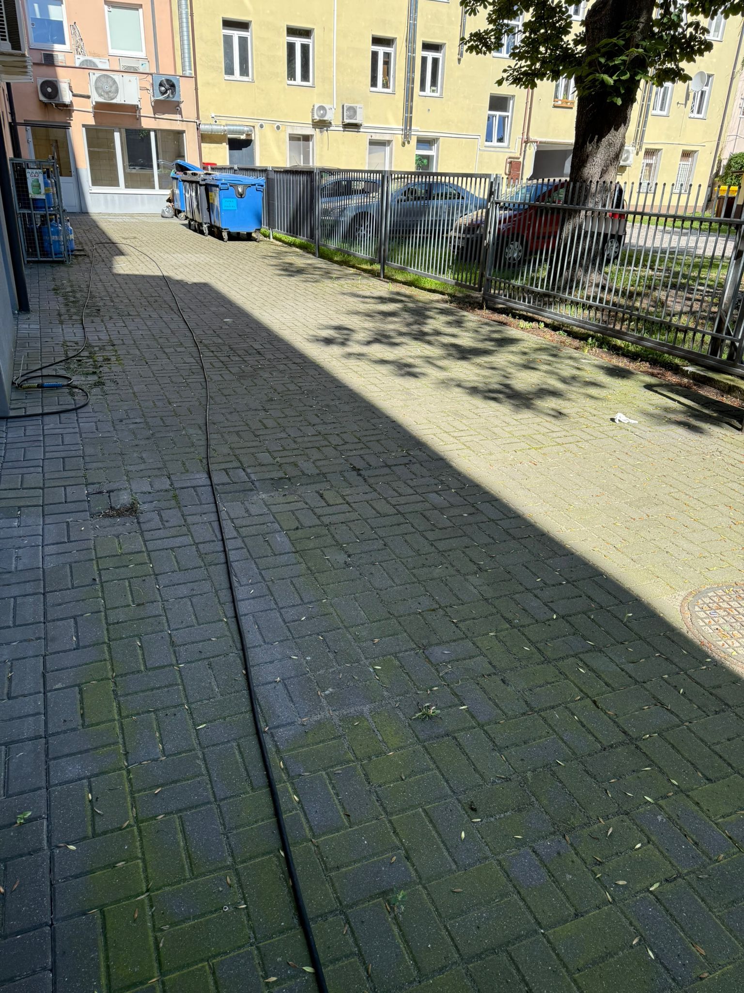 Ciemna kostka brukowa częściowo pokryta mchem, z kablem i sprzętem do czyszczenia w tle, sugerująca przygotowania do mycia. Budynek i drzewo w tle.