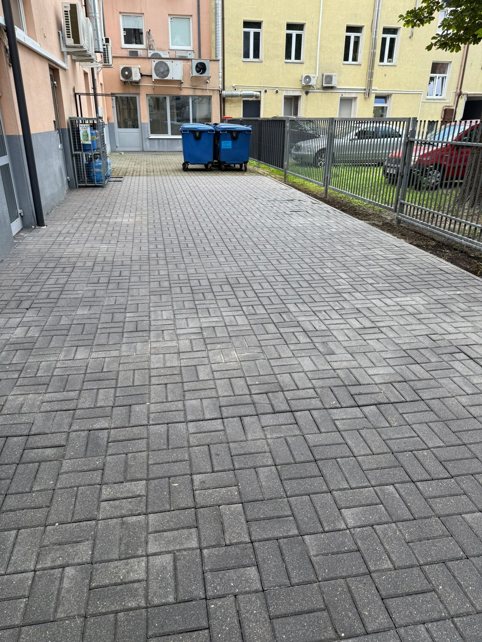 Szara kostka brukowa ułożona w wzór jodełki, z widocznymi budynkami w tle i metalowym ogrodzeniem. Dwa niebieskie pojemniki na śmieci w oddali.