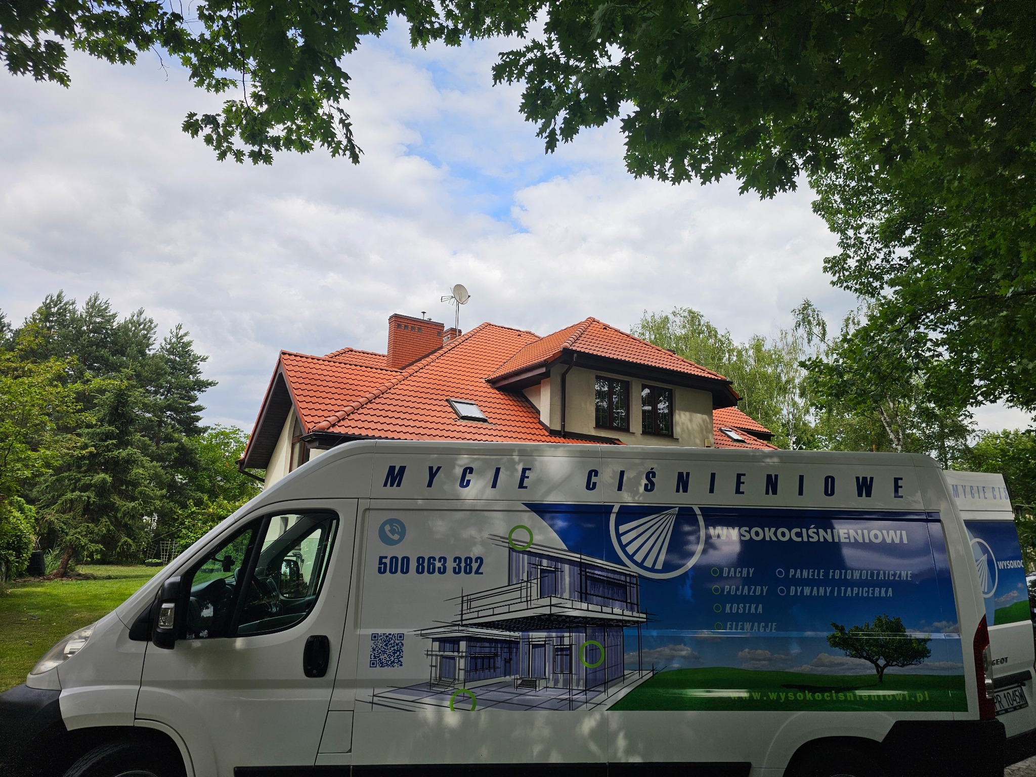 Biały van z reklamą mycia ciśnieniowego na tle domu z czerwoną dachówką i zieleni. Na vanie numer telefonu i lista usług: dachy, elewacje, kostka.