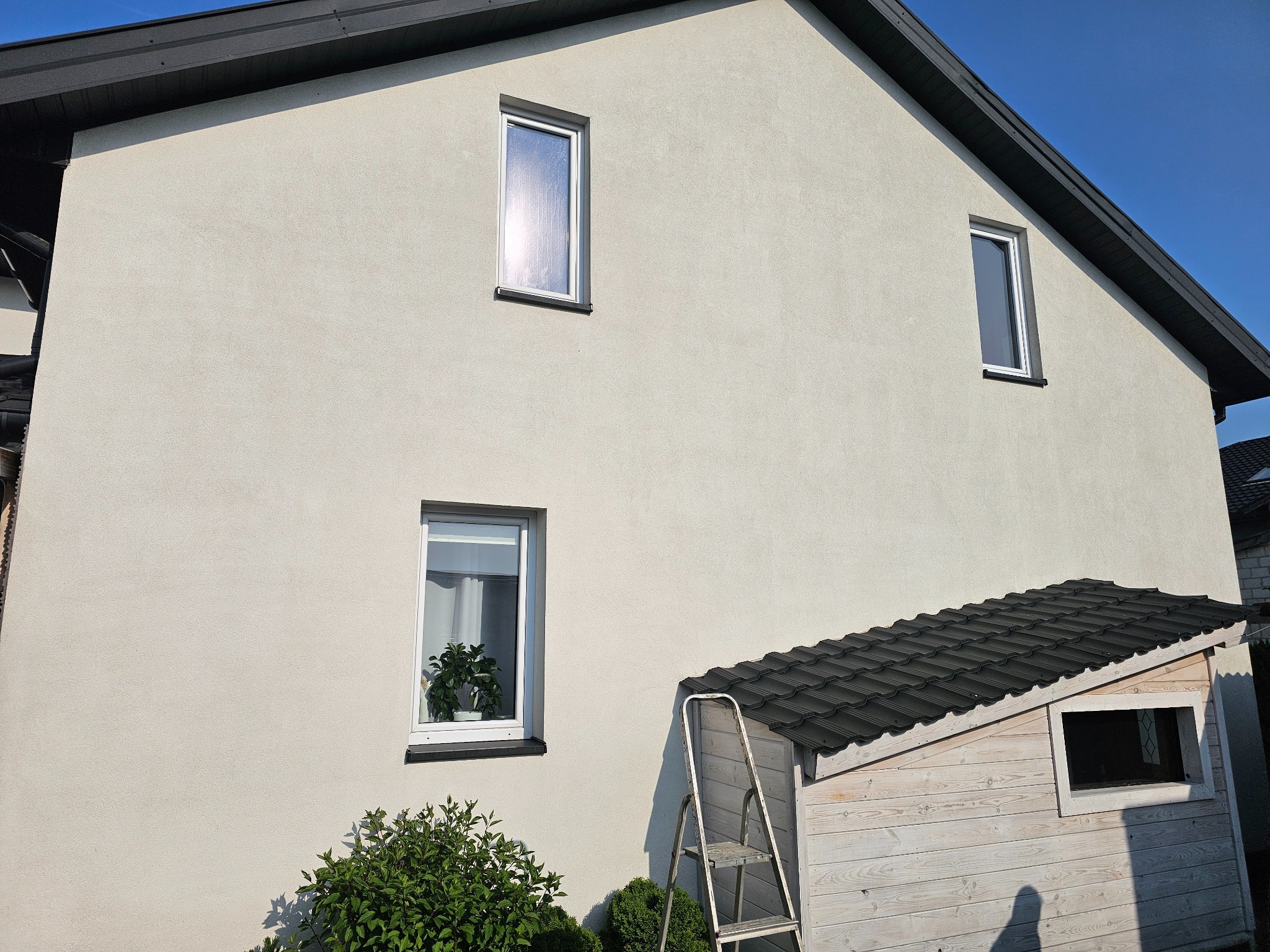 Jasna elewacja domu z trzema oknami, drabina oparta o drewnianą szopę z czarnym dachem. Krzew przed ścianą. Błękitne niebo w tle.