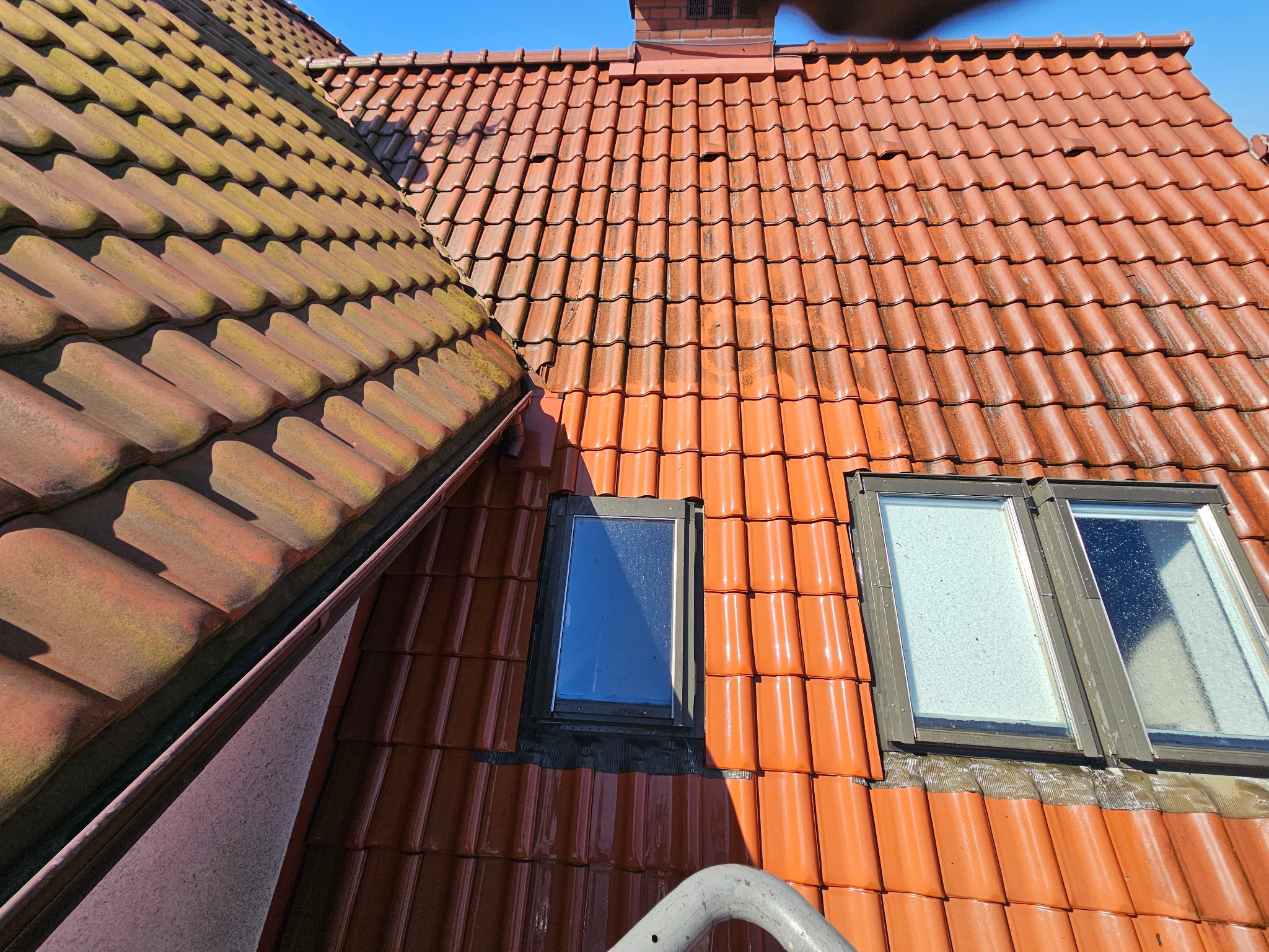 Czerwona dachówka po umyciu, widoczne okna dachowe i fragment drabiny. Część dachu po lewej stronie jest jeszcze zabrudzona, co uwidacznia efekt czyszczenia.