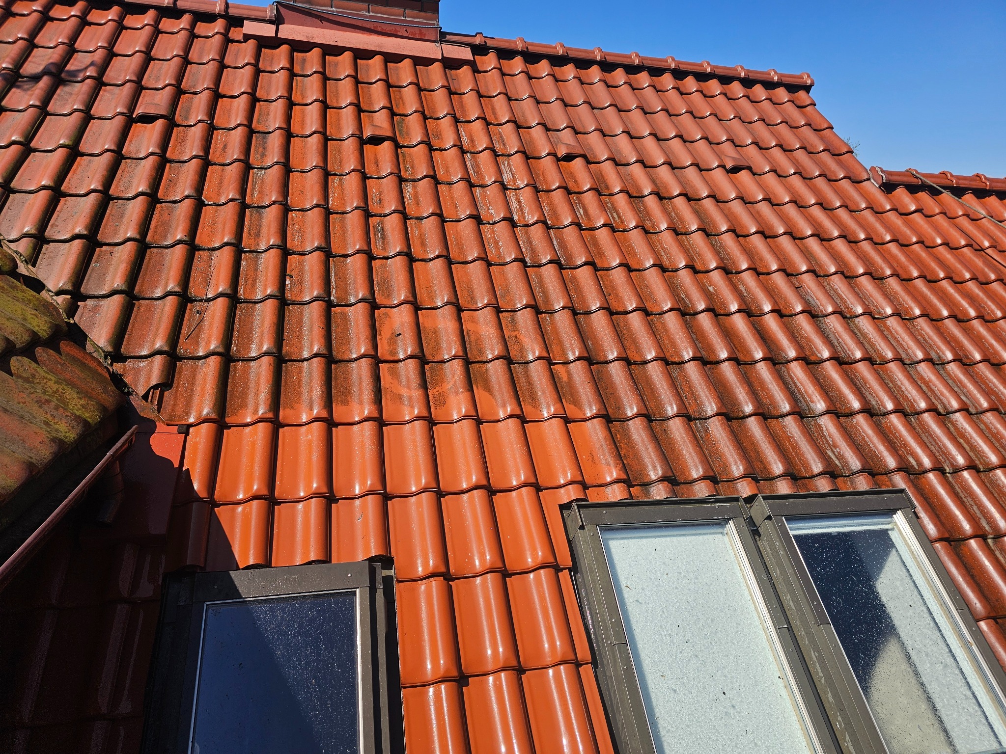Czerwona dachówka karpiówka, częściowo pokryta mchem, z oknami dachowymi. Widok z góry, w słoneczny dzień. Fragment komina w tle.