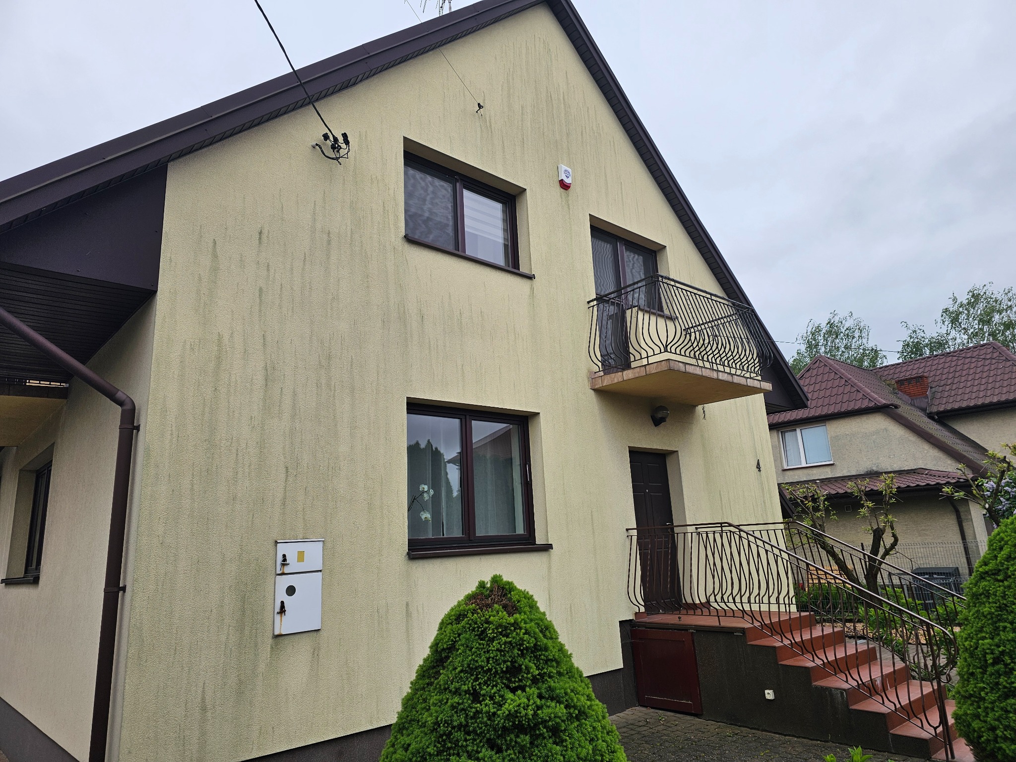Żółta elewacja domu z widocznymi zabrudzeniami i zaciekami, brązowe okna i dach, balkon z ozdobną balustradą. Widok z poziomu gruntu.