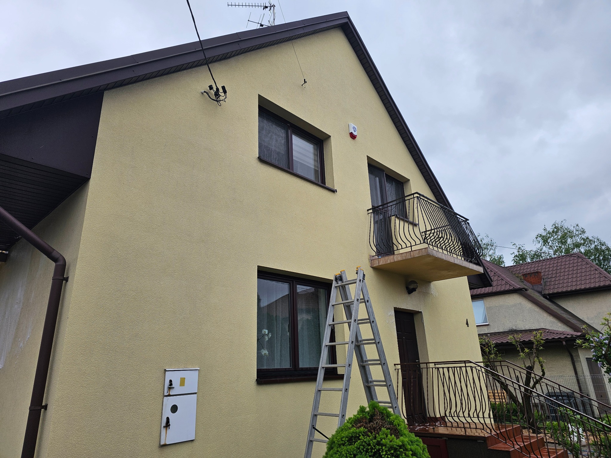 Dwupiętrowy dom z beżową elewacją, balkonem i drabiną opartą o ścianę. Widoczne okna z ciemnymi ramami, brązowe rynny i fragment dachu sąsiedniego budynku.