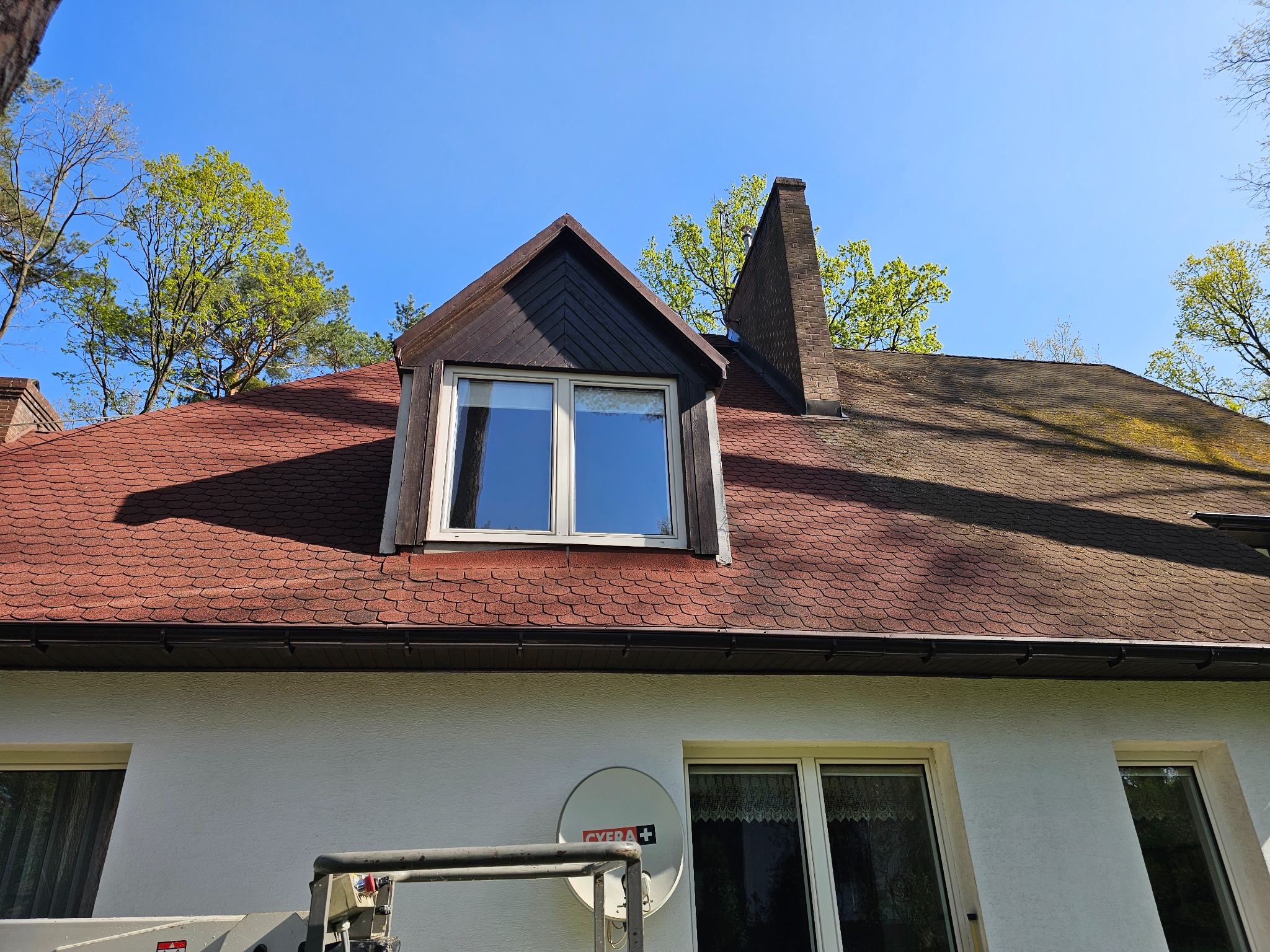 Czerwony dach z bitumicznej dachówki, komin i okno dachowe. Część elewacji z oknami i anteną satelitarną. Widok z podnośnika koszowego.