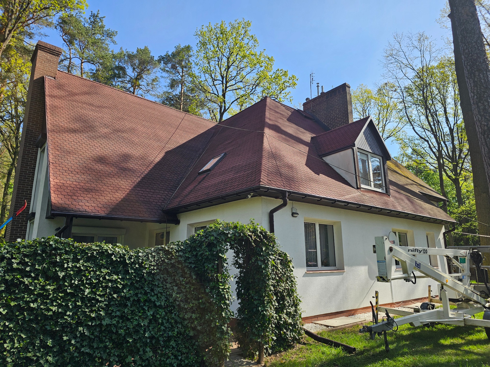 Dom z czerwoną dachówką i białą elewacją, widoczny podnośnik koszowy. Ujęcie z poziomu gruntu, zieleń w tle. Słoneczny dzień.