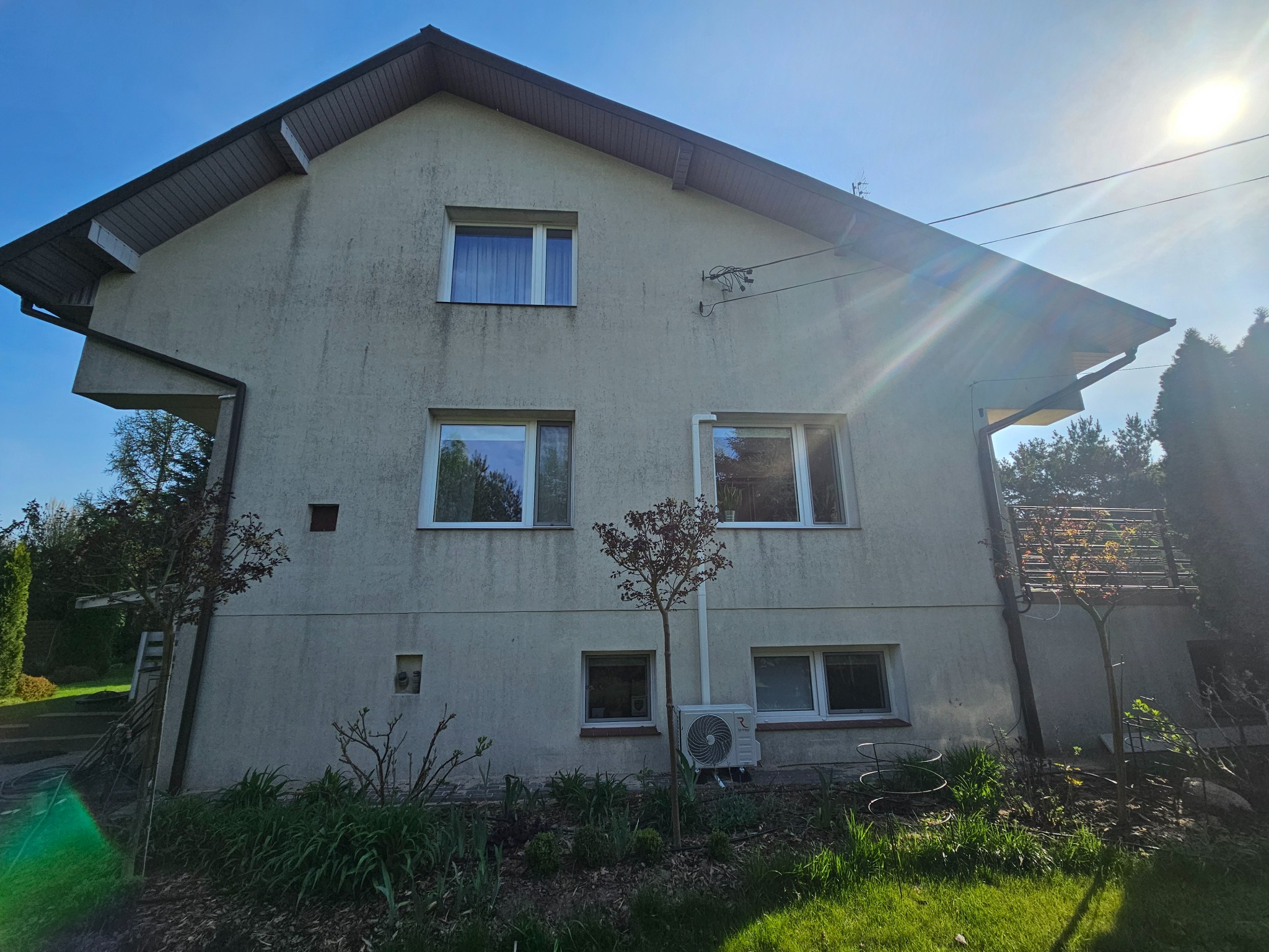 Dwupiętrowy dom z jasną elewacją w słoneczny dzień. Widoczne okna, klimatyzator na ścianie i zieleń w ogrodzie. Dom przed czyszczeniem elewacji.
