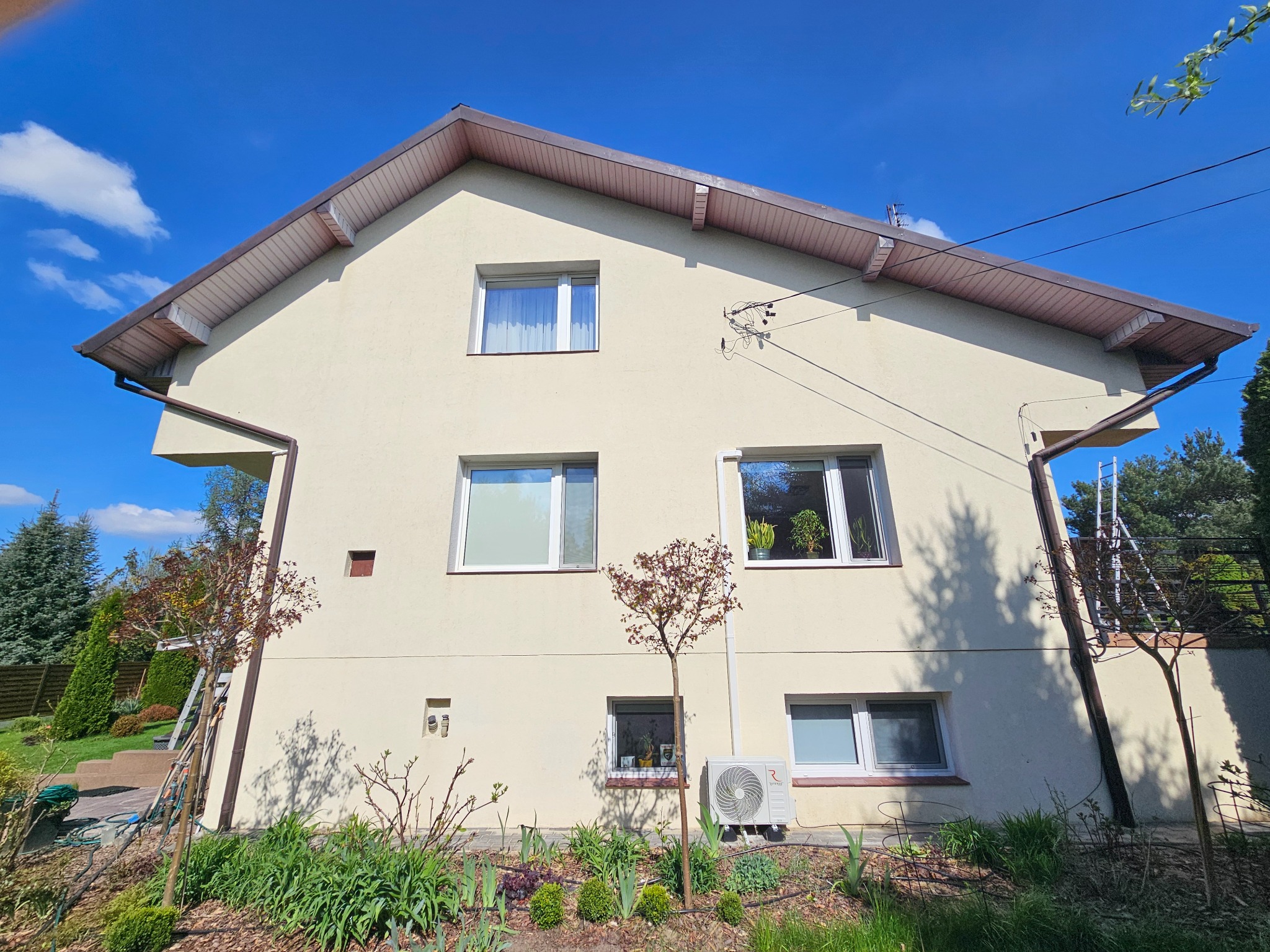 Dwupiętrowy dom z beżową elewacją w słoneczny dzień. Widoczne okna, rynny, drzewa i trawnik. Na ścianie zamontowany klimatyzator. Drabina oparta o ścianę.