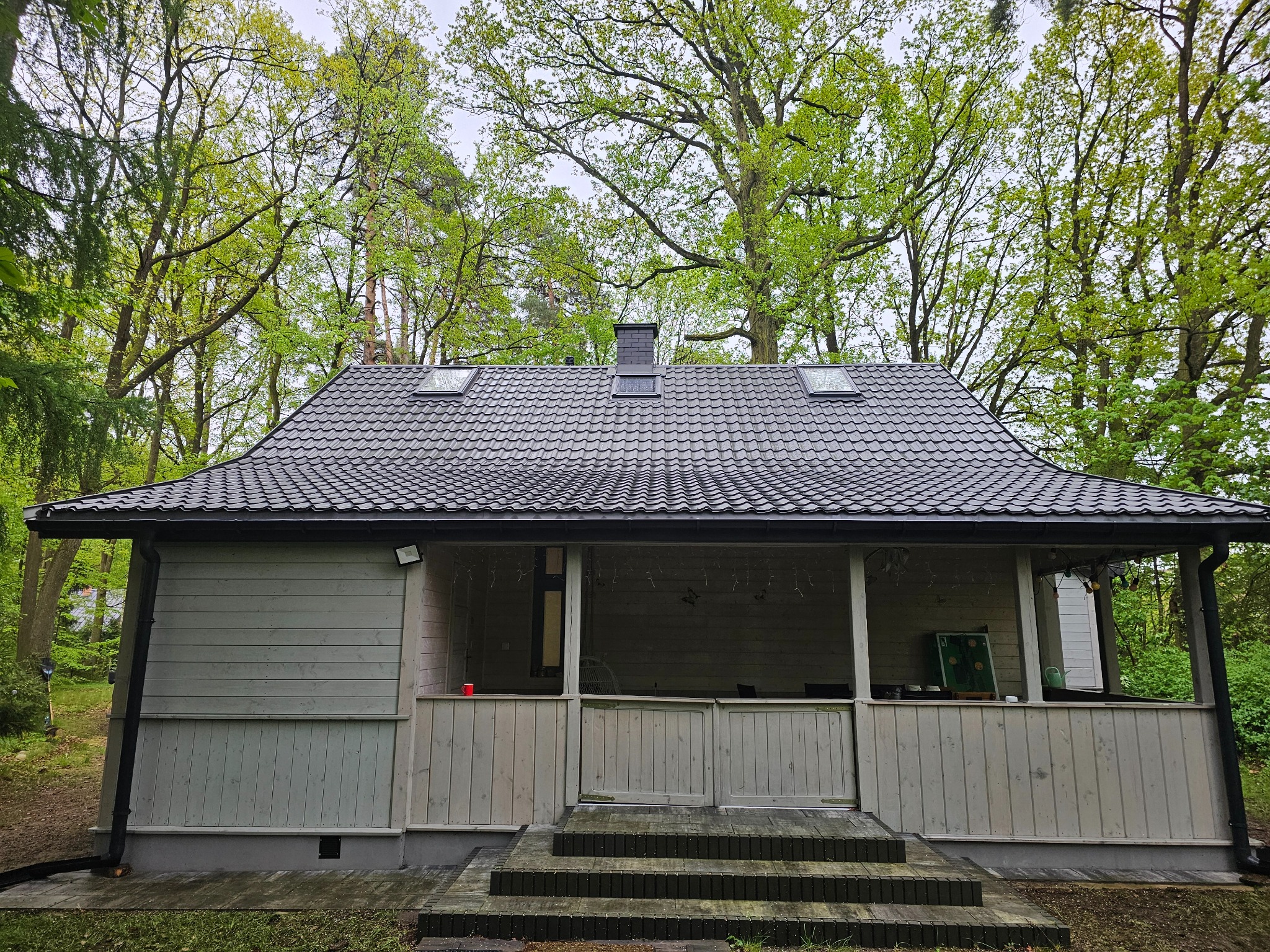 Szary, drewniany domek letniskowy z ciemnym dachem na tle zielonych drzew. Widoczne okna dachowe i schody wejściowe. Ujęcie z poziomu gruntu.