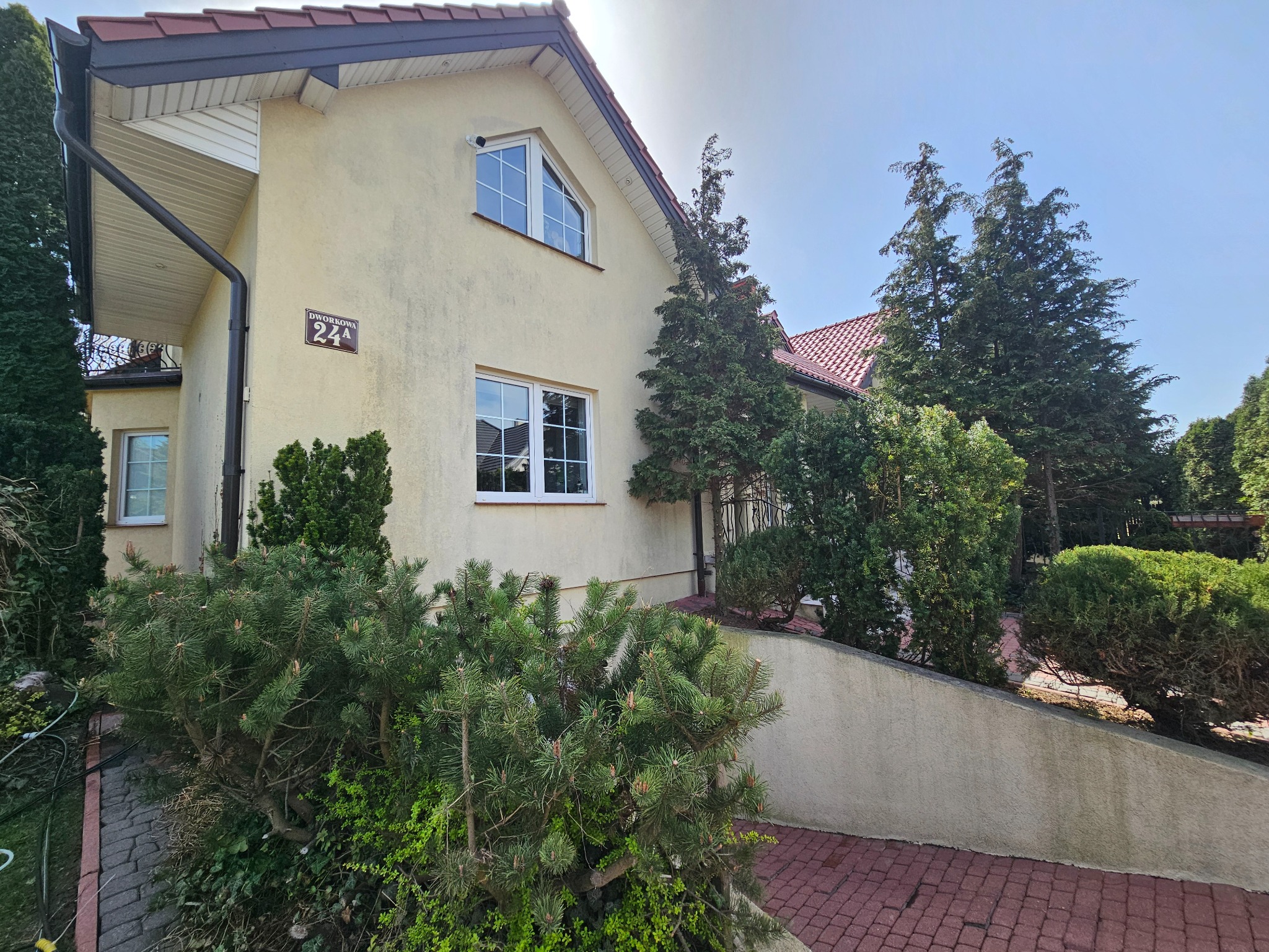 Dwupiętrowy dom z beżową elewacją, czerwonym dachem i brązowymi rynnami. Widoczne okna z białymi ramami, tabliczka z adresem i zadbana zieleń wokół budynku.