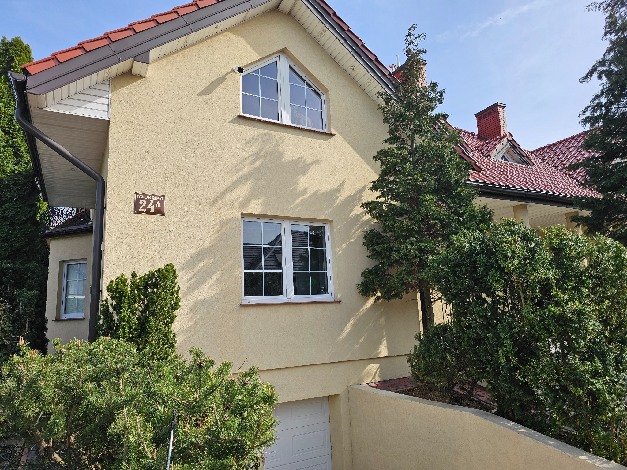 Jasna elewacja domu z beżowym tynkiem, widoczne okna z białymi ramami, brązowy dach i zieleń drzew. Tabliczka z adresem Dworkowa 24A.