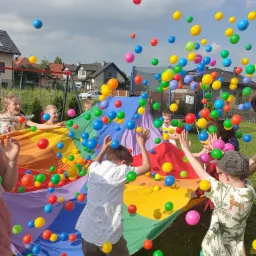 Grupa dzieci bawi się kolorową chustą animacyjną, podrzucając w górę mnóstwo kolorowych plastikowych piłek na tle domów i drzew.