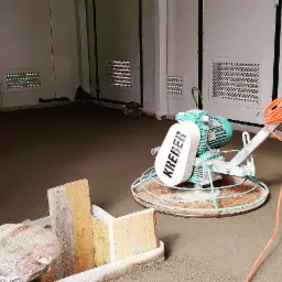 Zacieraczka mechaniczna marki Kreber na świeżo wylanej posadzce w pomieszczeniu z białymi ścianami i kratkami wentylacyjnymi.