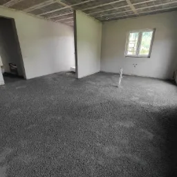 Wnętrze domu w stanie surowym, z wylaną, świeżą, ciemnoszarą posadzką jastrychową, widoczne okno, ściany i sufit w szarym tynku.