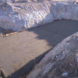 Świeżo wylana, gładka, szara płyta betonowa w wykopie ziemnym, widoczne ślady po narzędziach do rozprowadzania betonu i drewniane kołki stabilizujące.