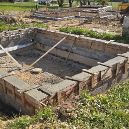 MK.Posadzki - Budowa fundamentów: wylany beton w szalunku, widoczny wykop i zbrojenie. Na drugim planie ciężarówka i prace ziemne.