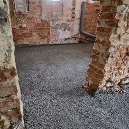 MK.Posadzki - Wnętrze w trakcie remontu z wylaną, szarą posadzką. Widoczne fragmenty ceglanych ścian, instalacja elektryczna i rury. Okno w tle.