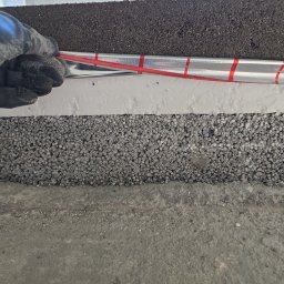 MK.Posadzki - Warstwy izolacyjne pod wylewkę: styropian, folia aluminiowa z czerwoną taśmą, czarna mata. Ręka w rękawicy układa instalację. Detal z budowy posadzki.