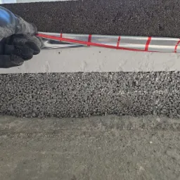 Warstwy izolacyjne pod wylewkę: styropian, folia aluminiowa z czerwoną taśmą, czarna mata. Ręka w rękawicy układa instalację. Detal z budowy posadzki.