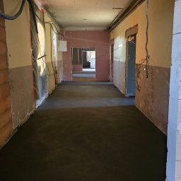 Wnętrze w trakcie remontu z nową, ciemną wylewką podłogową. Widoczne ściany w stanie surowym, instalacje i przejścia do kolejnych pomieszczeń. Perspektywa długa, głębia ostrości.