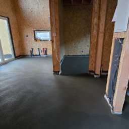 Świeżo wylana, gładka posadzka w surowym wnętrzu domu z drewnianą konstrukcją ścian. Widoczne instalacje wodne i elektryczne. Minimalistyczny, nowoczesny styl.