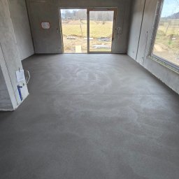 MK.Posadzki - Świeża, gładka wylewka w surowym wnętrzu z oknami na zewnątrz. Sufit zabezpieczony folią. Widoczne instalacje na ścianach. Naturalne światło dzienne.