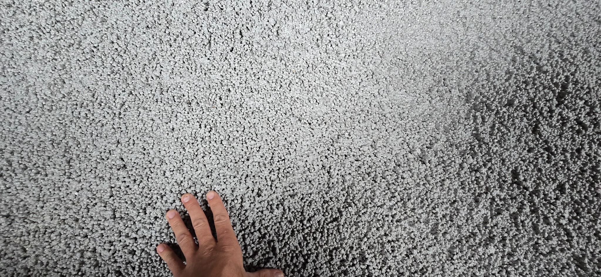 Szara, świeżo wylana posadzka jastrychowa o ziarnistej strukturze. W lewym dolnym rogu widoczna dłoń, prawdopodobnie pracownika, dotykająca powierzchni.