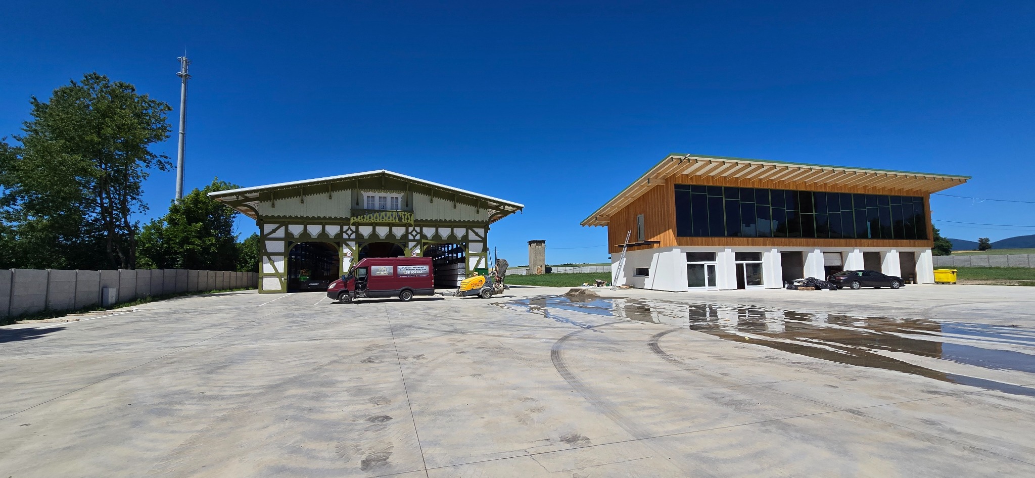 Rozległy plac z betonową nawierzchnią, z dwiema konstrukcjami budowlanymi w tle. Widoczny samochód dostawczy i maszyna do obróbki betonu. Błękitne niebo.