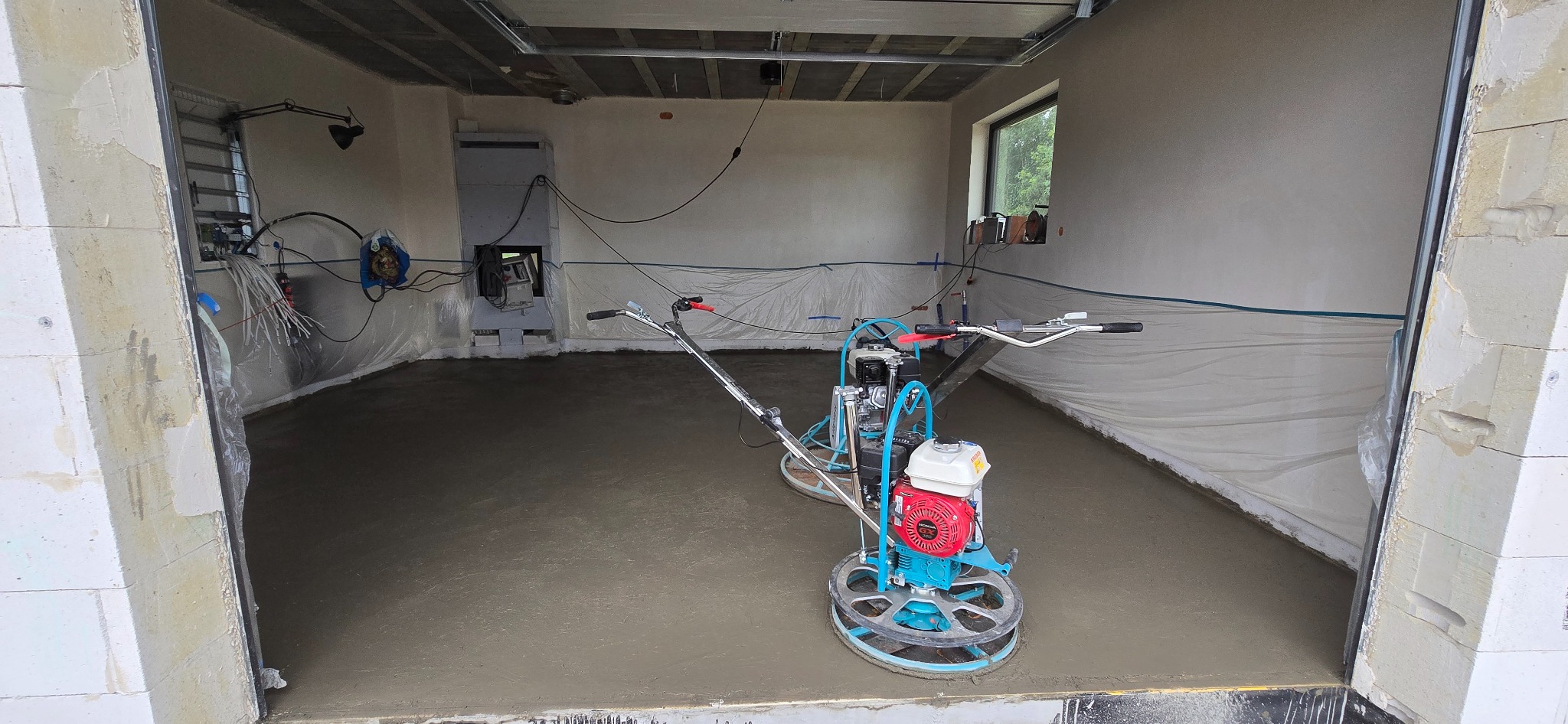 Dwie zacieraczki mechaniczne na świeżo wylanej, mokrej posadzce w surowym wnętrzu budynku. Folia ochronna przy ścianach, widoczne okno i instalacje.