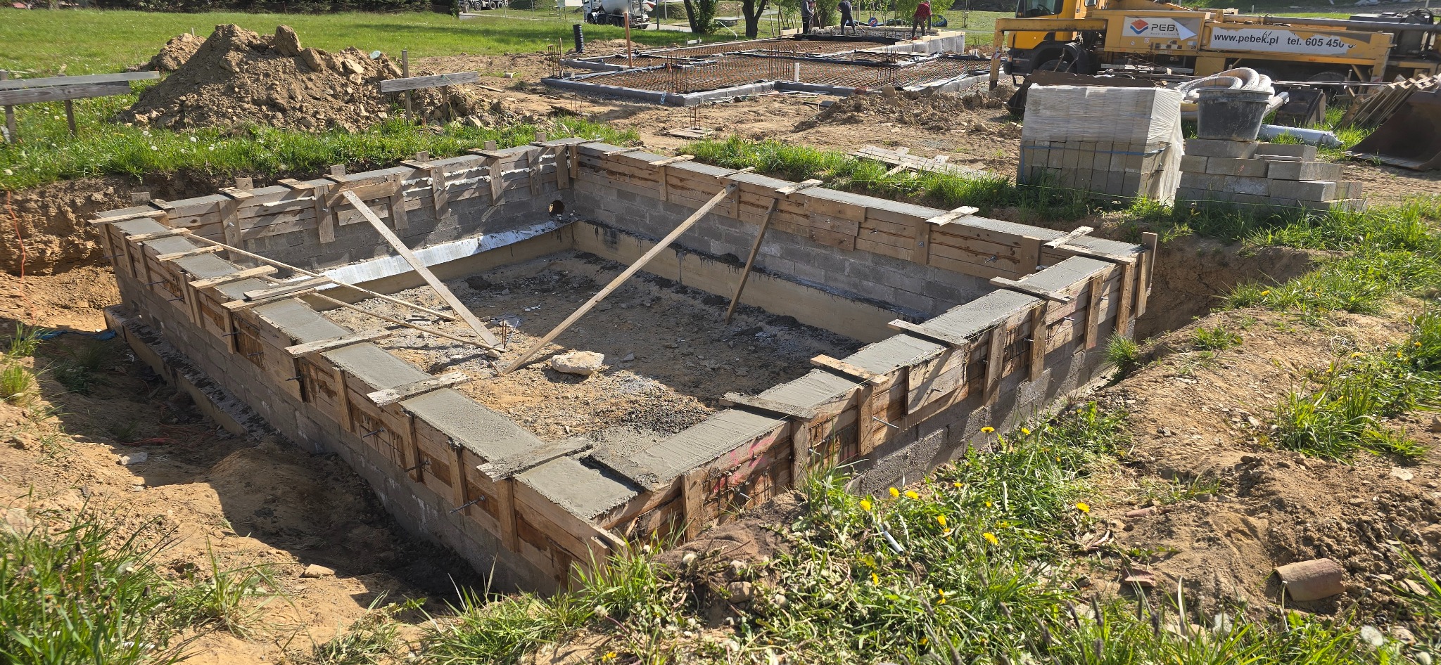 Budowa fundamentów: wylany beton w szalunku, widoczny wykop i zbrojenie. Na drugim planie ciężarówka i prace ziemne.
