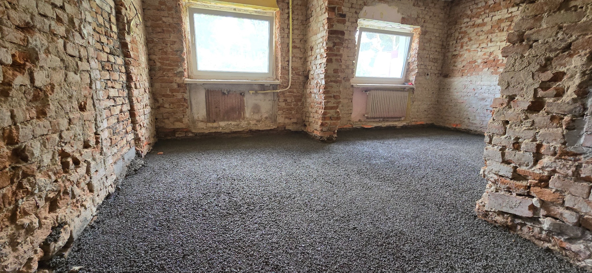Wnętrze z surowymi, ceglanymi ścianami i nową, szarą wylewką na podłodze. Dwa okna w tle wpuszczają naturalne światło. Widoczne fragmenty grzejnika i instalacji.