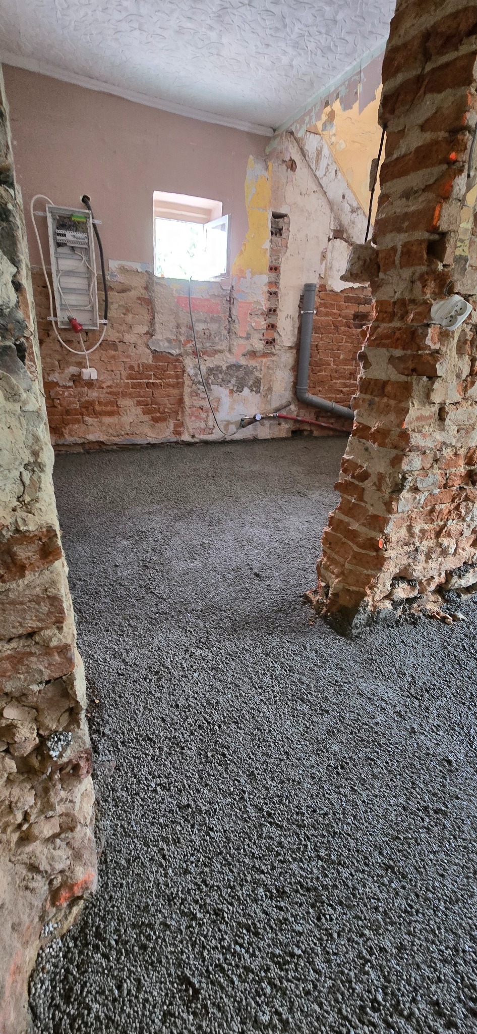 Wnętrze w trakcie remontu z wylaną, szarą posadzką. Widoczne fragmenty ceglanych ścian, instalacja elektryczna i rury. Okno w tle.