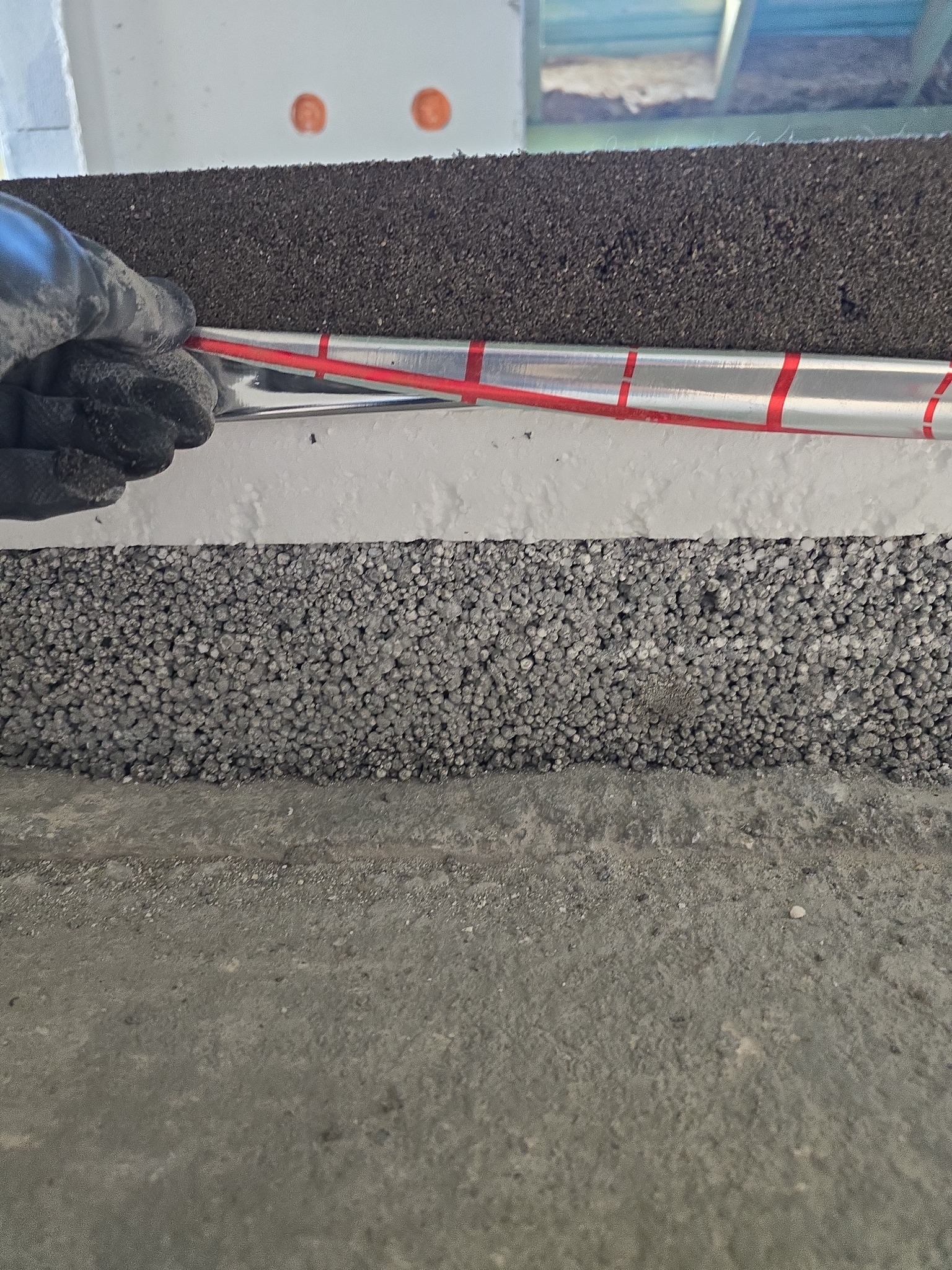 Warstwy izolacyjne pod wylewkę: styropian, folia aluminiowa z czerwoną taśmą, czarna mata. Ręka w rękawicy układa instalację. Detal z budowy posadzki.