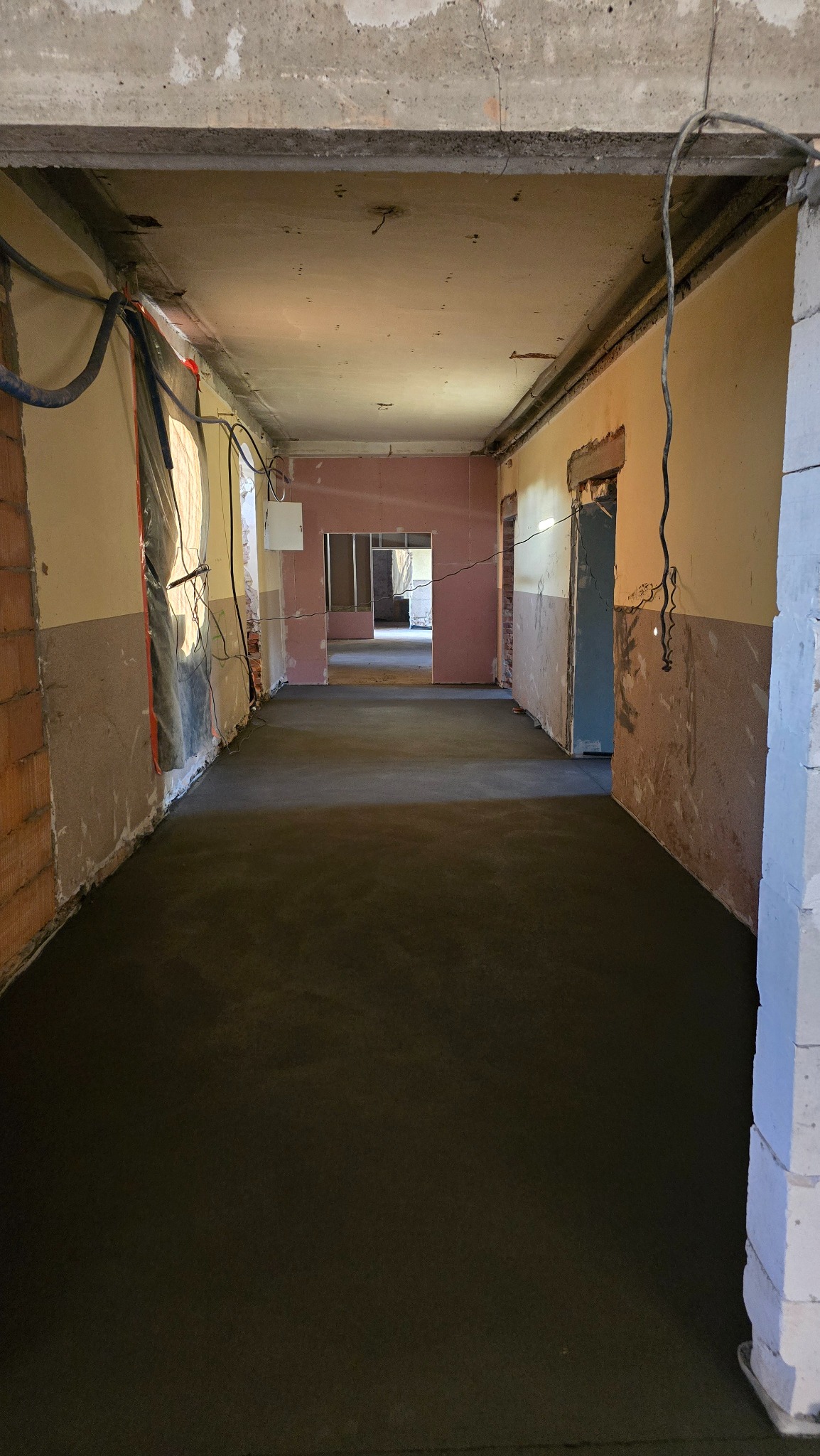 Wnętrze w trakcie remontu z nową, ciemną wylewką podłogową. Widoczne ściany w stanie surowym, instalacje i przejścia do kolejnych pomieszczeń. Perspektywa długa, głębia ostrości.