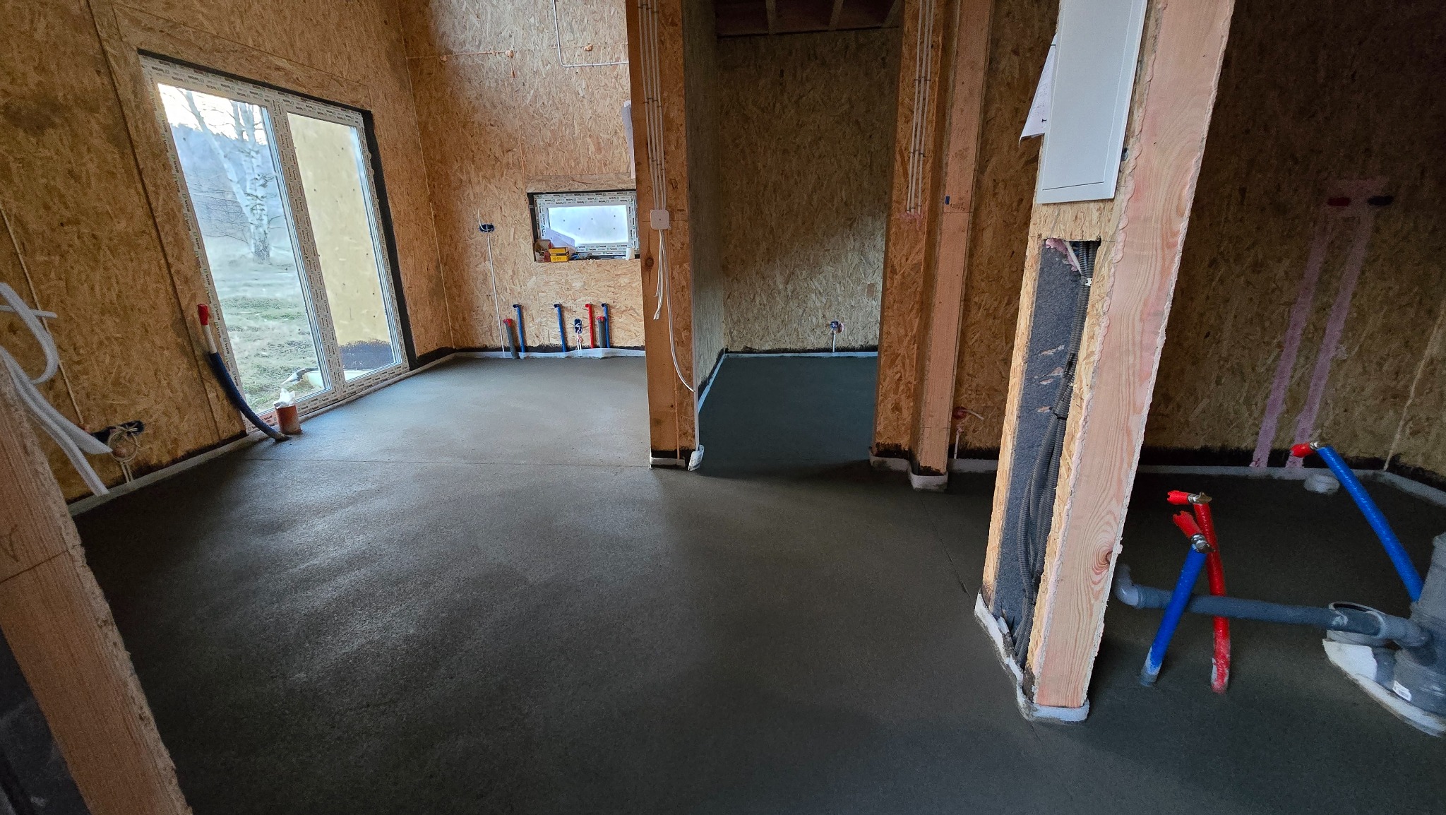 Świeżo wylana, gładka posadzka w surowym wnętrzu domu z drewnianą konstrukcją ścian. Widoczne instalacje wodne i elektryczne. Minimalistyczny, nowoczesny styl.