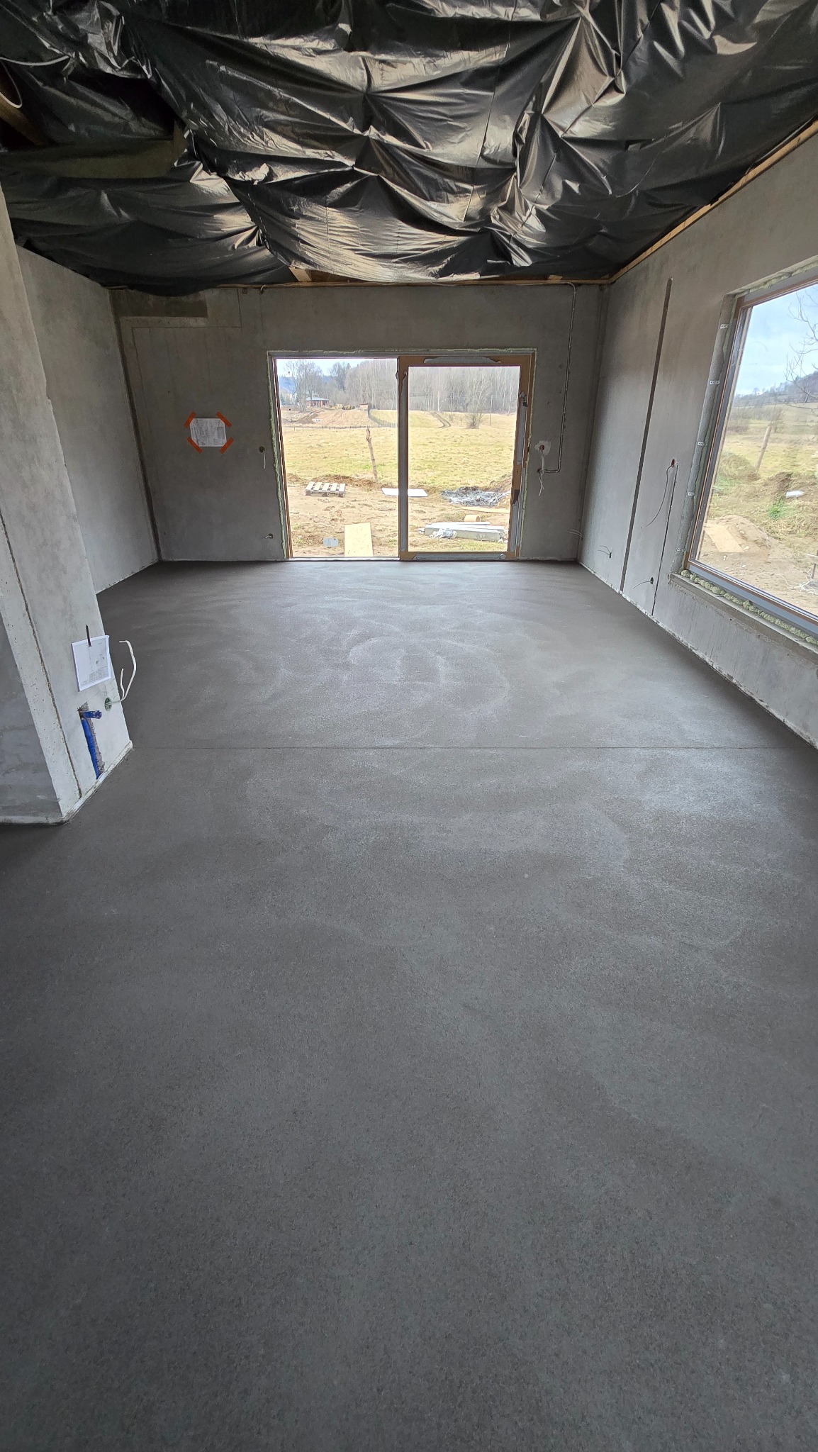 Świeża, gładka wylewka w surowym wnętrzu z oknami na zewnątrz. Sufit zabezpieczony folią. Widoczne instalacje na ścianach. Naturalne światło dzienne.
