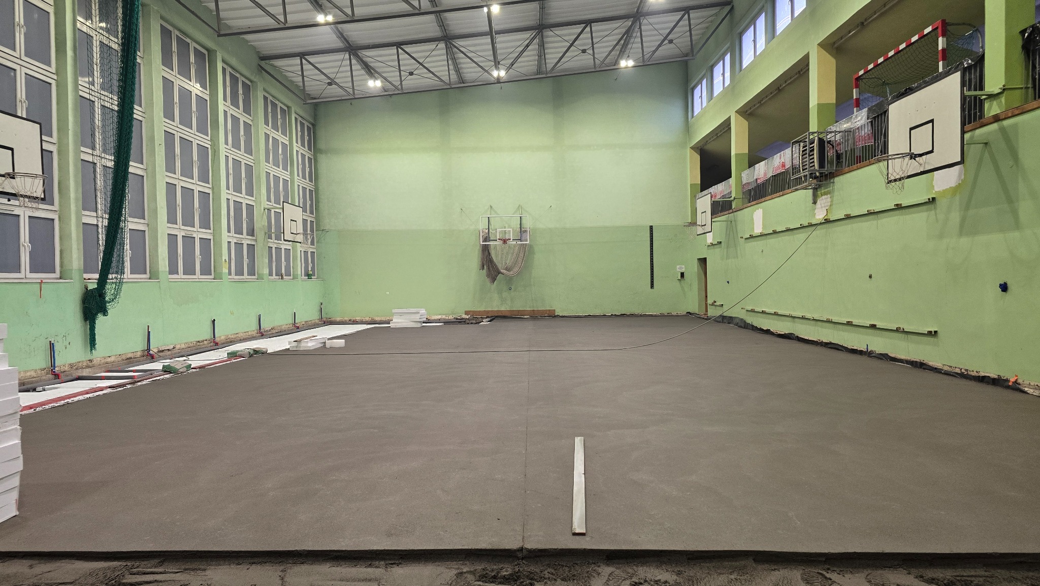 Wnętrze sali gimnastycznej z nową, szarą wylewką podłogową. Widoczne kosze do koszykówki na ścianach i materiały izolacyjne przygotowane do dalszych prac.